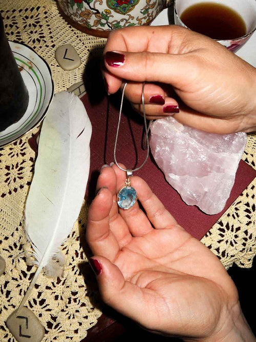 Person holding a necklace with a blue pendant on a table with a feather and crystal.
