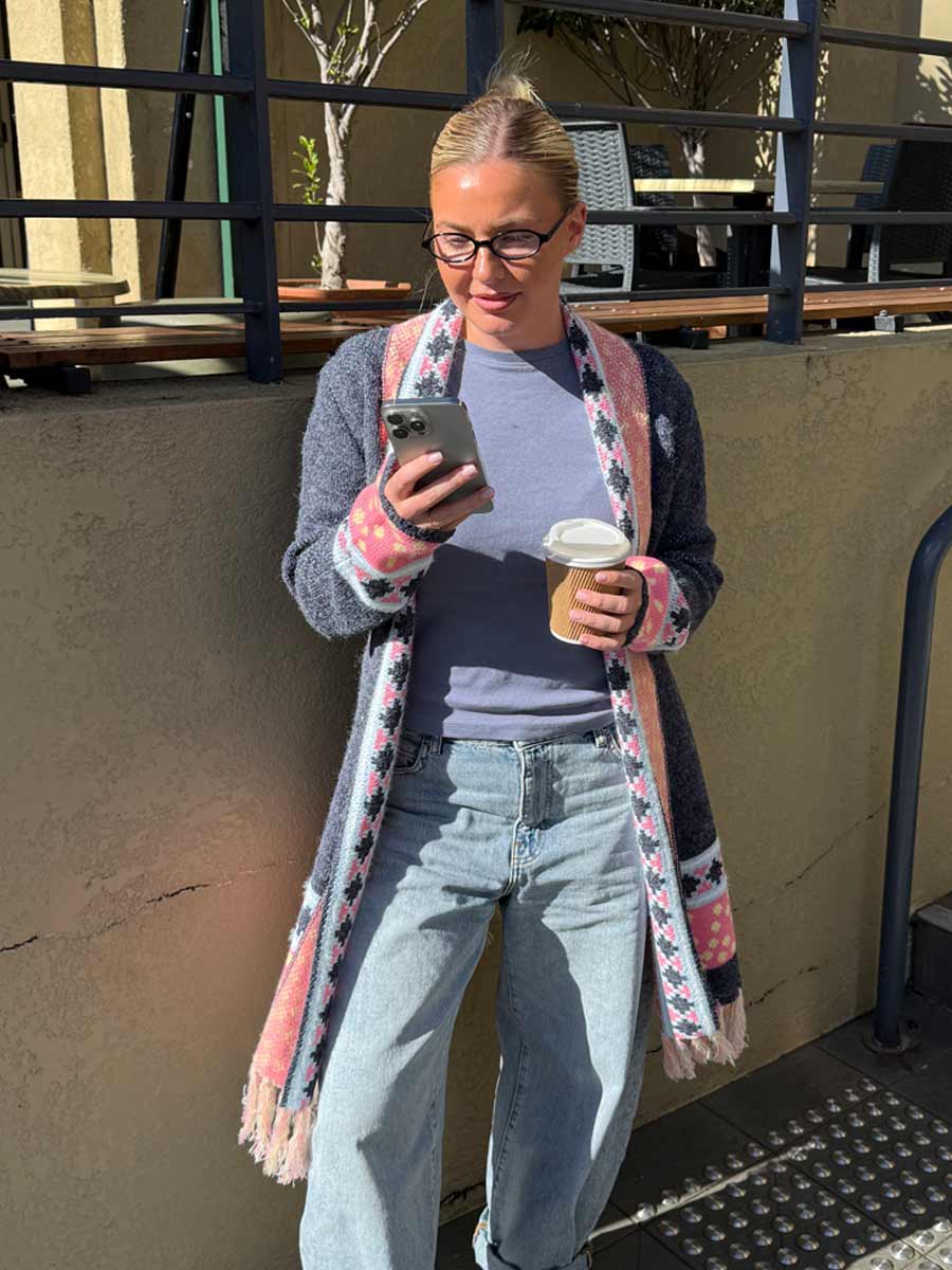A woman wearing a gray knit cardigan with pink and white geometric patterns, a light blue t-shirt, and light-wash jeans, holding a coffee cup and looking at her phone outdoors.