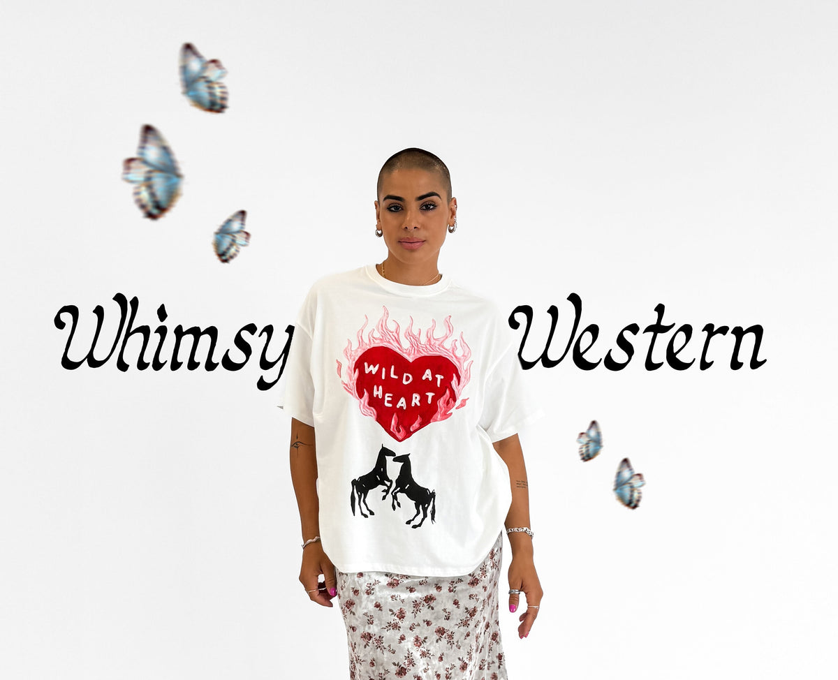 A model stands against a white studio background wearing a white graphic t-shirt with a red heart and black horse design, paired with a floral midi skirt and black boots. The text 'Whimsy Western' is written on the left side of the image with decorative butterflies.