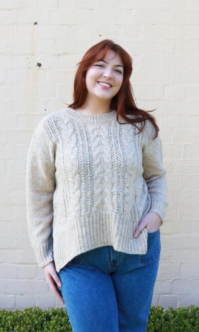 A woman wearing a beige, cable knit sweater with side splits.