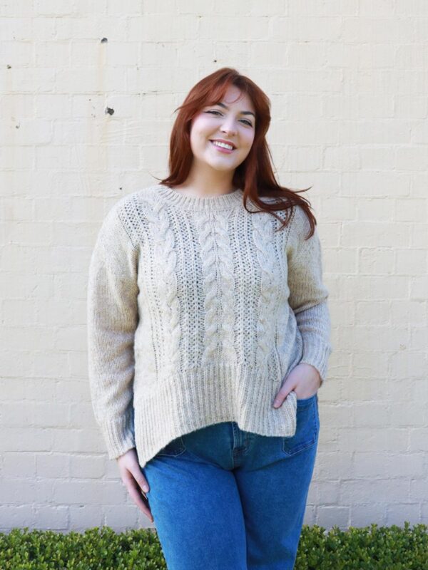 A woman wearing a beige, cable knit sweater with side splits.