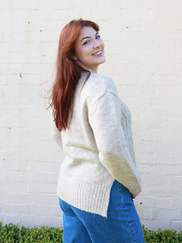 A woman wearing a beige, cable knit sweater with side splits.