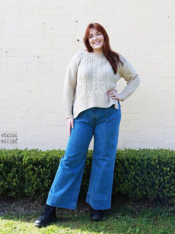 A woman wearing a beige, cable knit sweater with side splits.