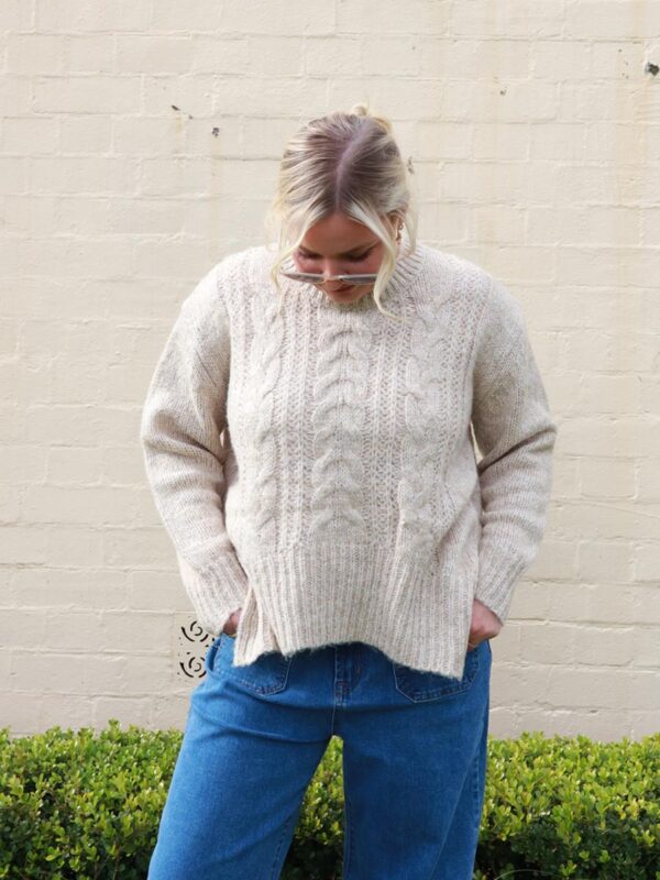 A woman wearing a beige, cable knit sweater with side splits.