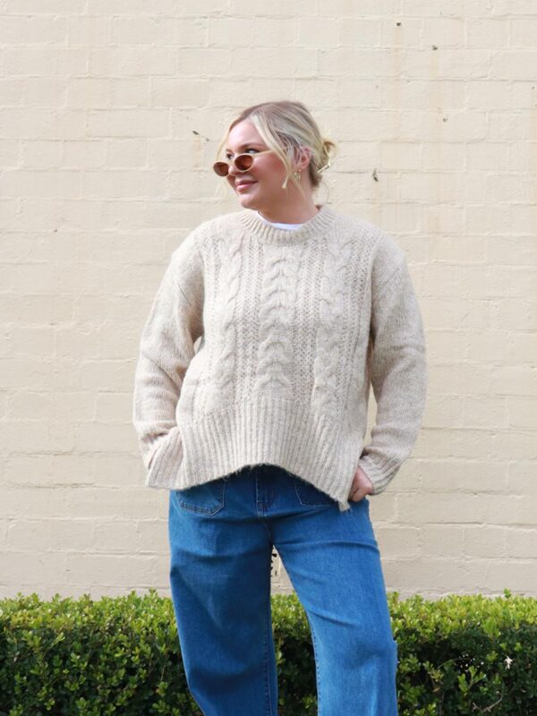 A woman wearing a beige, cable knit sweater with side splits.