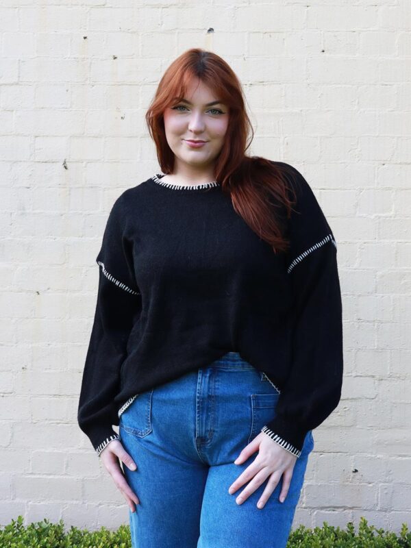 A woman wearing a black knit sweater with white contrast stitching on the cuffs and hems.