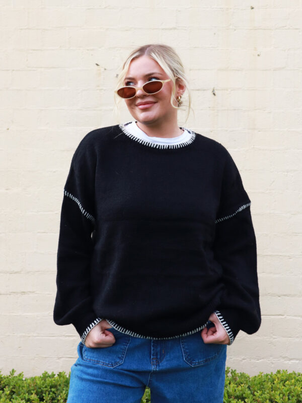 A woman wearing a black knit sweater with white contrast stitching on the cuffs and hems.